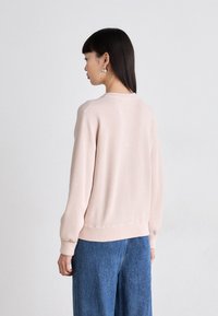 Pull rose clair avec un col rond et des manches longues, doté de poignets et d'un ourlet côtelés. Porté avec un jean bleu à jambes larges, au design minimaliste.