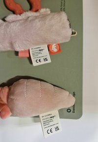 Two soft plush toys in shades of pink, featuring a textured design with labels attached. Background includes a green mat with manufacturer details.