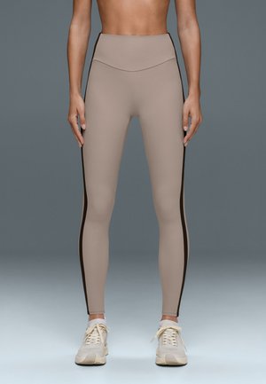 Femme portant un legging beige taille haute avec des bandes latérales noires et des chaussures de sport blanches, debout devant un fond gris.