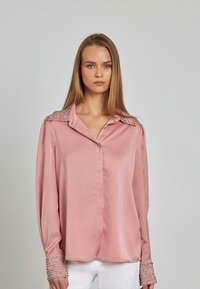 Langärmlige, hellrosa Satinbluse mit Kragen und Nietenakzenten an den Schultern sowie geknöpften Ärmeln, glatte Textur.