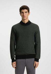 Dark green textured knit sweater with a round neckline and ribbed cuffs. Worn over a black collared shirt, paired with light gray pants.
