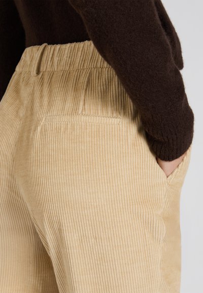 Pantalones de pana en beige con canales verticales. Cintura elástica y bolsillos laterales. Prenda marrón parcialmente visible en la parte superior.