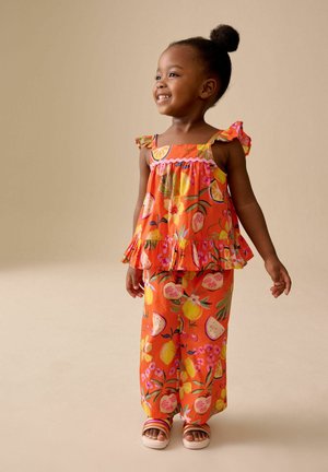Lächendes junges Mädchen in einem leuchtend orangefarbenen, ärmellosen Oberteil und Hosen mit tropischem Fruchtmuster, stehend vor einem einfarbigen beigefarbenen Hintergrund.