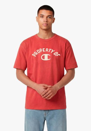 Giovane uomo che indossa una maglietta rossa con la scritta "PROPERTY OF" e un logo, in piedi con le mani leggermente intrecciate, jeans azzurri, sfondo neutro.