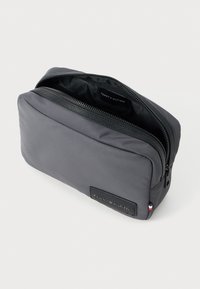 Šedá toaletní taška z látky s uzávěrem na zip, černými akcenty a logem. Hladký povrch, obdélníkový tvar a minimalistický design.