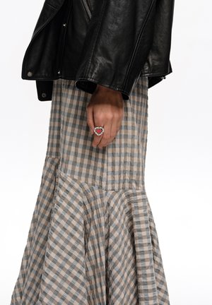 Ręka spoczywająca obok ciała, nosząca pierścionek w kształcie serca; strój obejmuje czarną skórzaną kurtkę i długą beżowo-szaro kratowaną spódnicę.