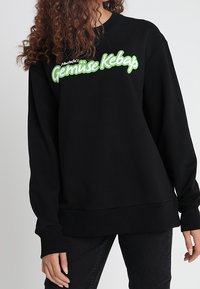 Black sweatshirt with green text reading "Gemüse Kebap." Features a relaxed fit, ribbed cuffs, and hem. Soft fabric texture.