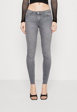 Slim fit grijze jeans gedragen met een zwart visnet lange mouw top en zwarte open schoenen, die de buik en benen tonen tegen een witte achtergrond.