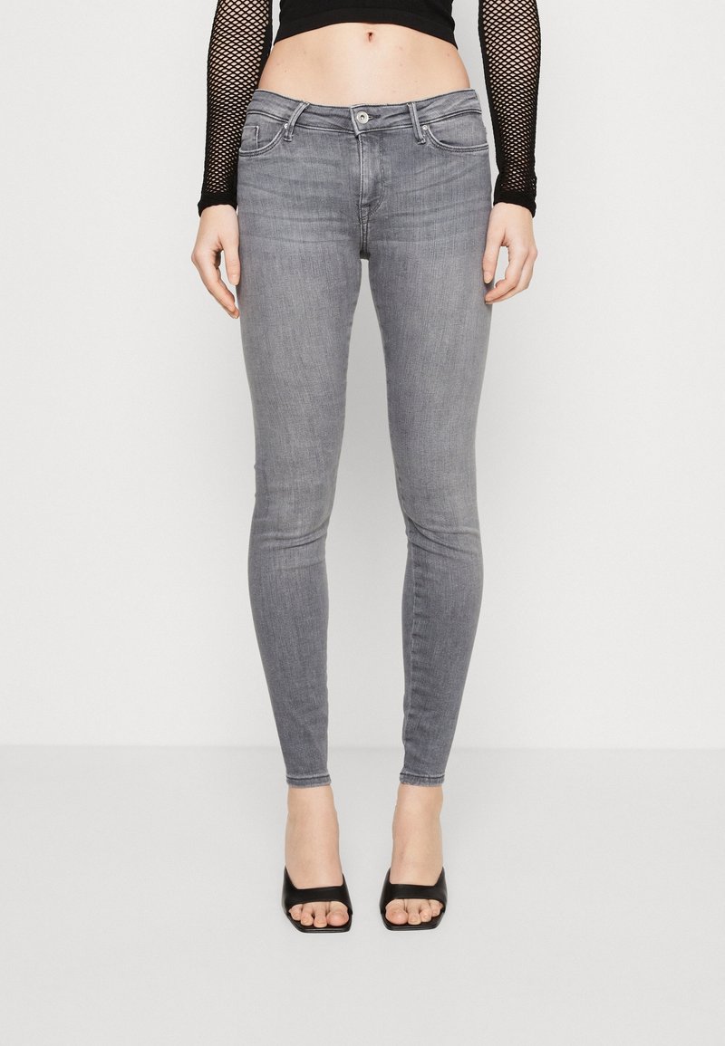 Jean gris slim porté avec un top noir en résille à manches longues et des talons ouverts noirs, montrant le ventre et les jambes sur fond blanc.