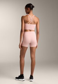 Svetloružový športový podprsenka s prekríženými ramienkami, v kombinácii s vhodnými vysoko pásovými šortkami. Hladký, elastický materiál zdôrazňuje priliehavý strih.