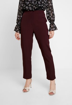 Kobieta w ciemnorubinowych prostych spodniach do kostek w połączeniu z beżowymi sandałami na wysokim obcasie oraz przezroczystą czarną bluzką w kwiatowy wzór z falbankami na mankietach.