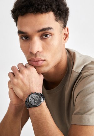 Junger Mann mit lockigem Haar, der ein beigefarbenes T-Shirt trägt und sein Kinn auf verschränkte Hände legt, wobei er eine schwarze Metallarmbanduhr trägt.