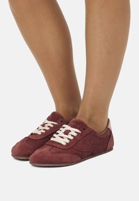 Sneakers in suede bordeaux con lacci bianchi, punta tondeggiante e dettagli in beige sul tallone. Presentano un design atletico classico e una superficie texture.