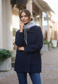 Marineblaue Wolljacke mit doppelreihiger Knopfleiste und seitlichen Taschen, über einem hellgrauen Kapuzenpullover getragen. Legere Jeans ergänzen das Outfit.