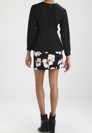 Kobieta stoi tyłem, ubrana w czarną bluzkę z długim rękawem, czarną mini spódnicę w kwiatowy wzór oraz czarne botki na obcasie.