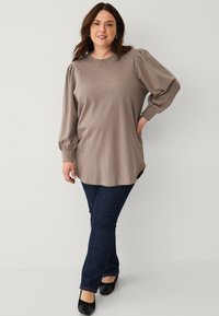 Long-sleeve taupe top with puffed shoulders, rounded neckline, and side slits, paired with dark blue fitted jeans and black block-heeled shoes.