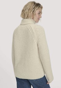 Cremfarbener, grob gestrickter Pullover mit hohem Kragen, geripptem Textur und überschnittenen Schultern. Lockerer Schnitt, sichtbare Nähte und seitliche Schlitze.