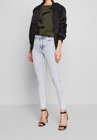 Černá bomber bunda, olivově zelené maskáčové tričko, světle modré skinny džíny a černé sandály na podpatku. Jednoduchý design, hladká textura.