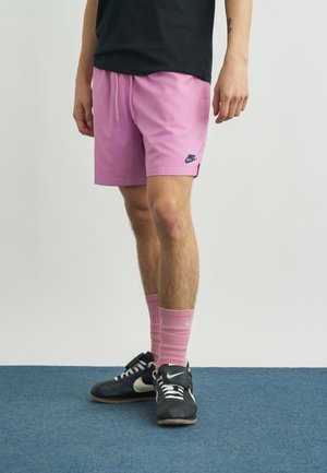 Personne portant un short Nike rose, des chaussettes Nike assorties roses, des baskets Nike noires, debout sur un tapis bleu devant un fond uni.