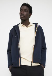 Marineblå zip-up hoodie med kontrasterende gule og røde striber, båret over en lys beige polo t-shirt, med en blød tekstur og afslappet pasform.