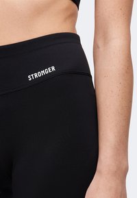 Primo piano di una persona che indossa leggings neri a vita alta con la parola "STRONGER" stampata vicino alla vita e parte del suo braccio visibile.