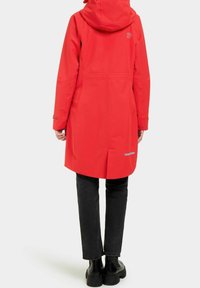 Röd vattentät rock med huva, slank design, lång modell och reflekterande detaljer. Bärs över svarta byxor och grova svarta stövlar.