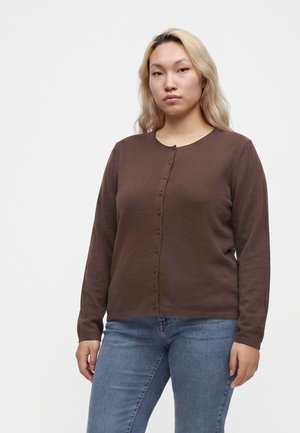 Ung person med langt blondt hår iført en brun knapcardigan og blå jeans mod en ensfarvet hvid baggrund.