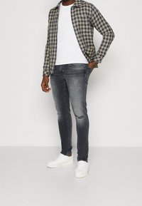 Kostkovaná košile v námořnické modři a žluté, bílé tričko, vyšisované šedé džíny s dírami a bílé tenisky. Model stojí s rukama v kapsách.