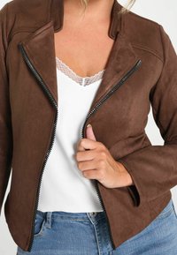 Ruskea mokkatakki, jossa on epäsymmetrinen vetoketjukiinnitys, tyylikäs muotoilu ja muotoiltu istuvuus, which on valkoisen pitsireunaisen topin päällä.