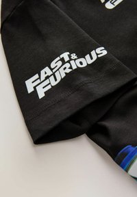 Mâneca din țesătură neagră cu text alb "FAST & FURIOUS" imprimat aproape de tiv și o mică fereastră a unei grafice colorate dedesubt.