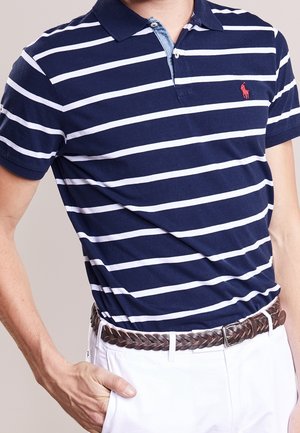 Marineblau- und weißgestreiftes Polohemd mit einem Denimkragen. Verfügt über kurze Ärmel und ein rotes Logo; kombiniert mit einem braunen geflochtenen Gürtel und weißen Hosen.