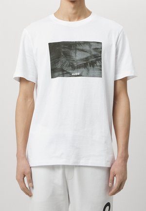 Mies, jolla on valkoinen T-paita mustalla suorakulmaisella kuviolla ja "HUGO"-teksti rinnan keskellä, yhdistettynä valkoisiin housuihin.