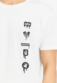 Tricou alb cu mânecă scurtă, cu imprimeuri grafice negre verticale, inclusiv simboluri pentru pace, libertate și o porumbel, cu un aspect uzat.