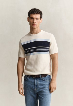 Homme portant un pull beige à manches courtes avec des rayures horizontales bleues et noires, un jean bleu et une ceinture marron, debout devant un fond uni.