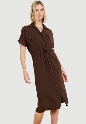 Blonde vrouw die een bruin midi-jurk met korte mouwen draagt, met knopen, borstzakken en een strikceintuur, staand tegen een lichte achtergrond.