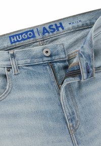 Jeans in denim azzurro chiaro con una finitura sbiadita, dotati di chiusura a bottone, patta con cerniera e cuciture a contrasto. L'etichetta del marchio legge "HUGO ASH."