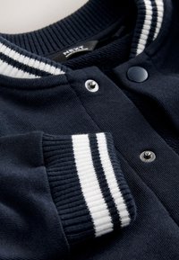 Gros plan sur une veste universitaire bleu marine avec col et poignets côtelés blancs rayés, et des boutons-pression en métal le long de l'ouverture avant.