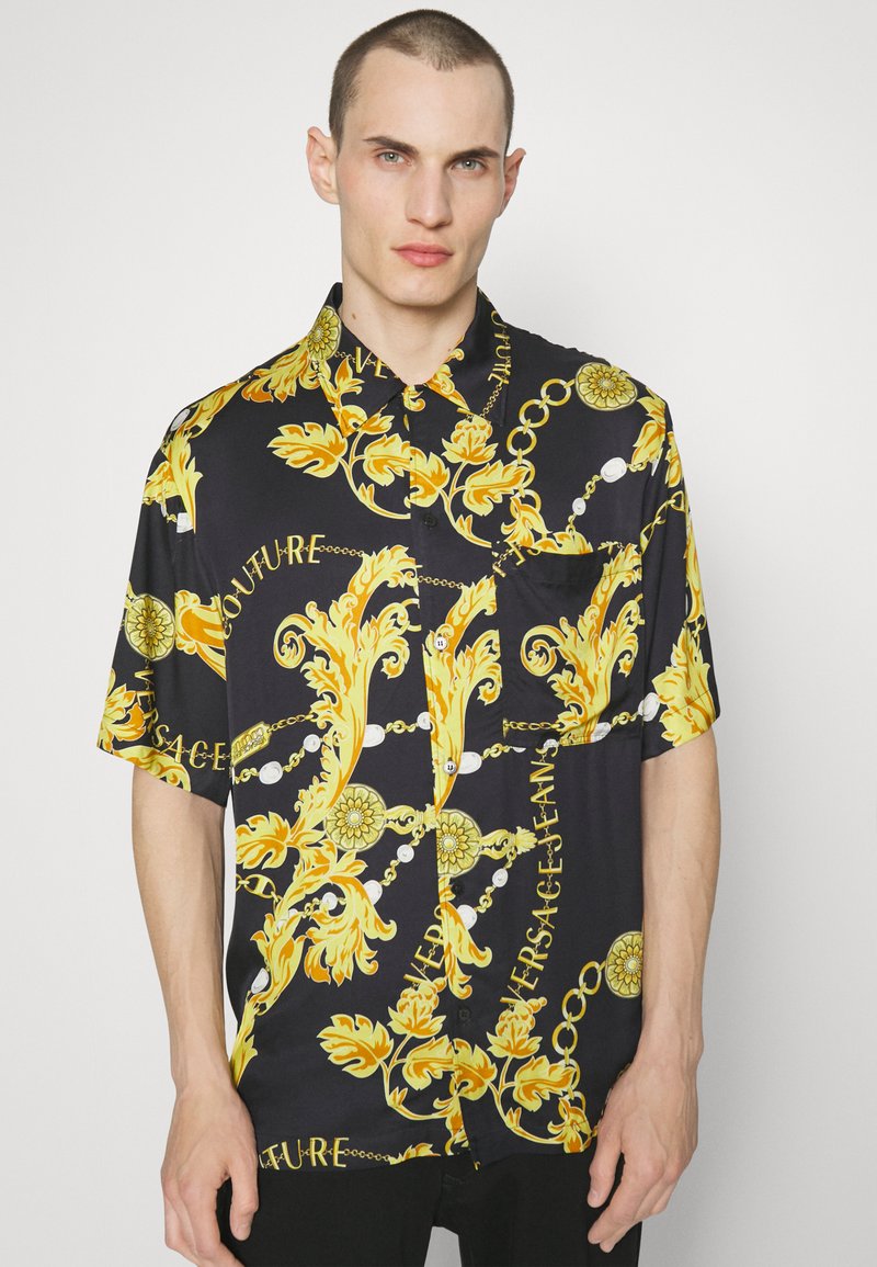 Chemise noire à manches courtes avec boutons, ornée d'un motif baroque jaune avec des chaînes et du texte. Comprend une poche sur la poitrine gauche. Tissu lisse.