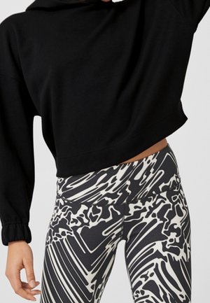 Persoon draagt een zwarte cropped hoodie en hooggetailleerde zwarte en witte legging met abstract patroon, staat met één arm omhoog.