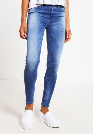 Jeans ajustados de mezclilla azul claro con un ligero desvanecimiento, cintura media y cremallera. Combinados con zapatillas blancas tipo slip-on.