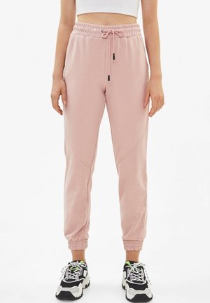 Persoon die lichtroze joggerbroek draagt met elastische taille en boorden, gecombineerd met zwart-witte chunky sneakers op een witte achtergrond.