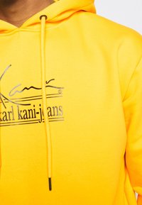 Sudadera amarilla de mezcla de algodón. Cuenta con capucha con cordón y un logo impreso en negro. Textura suave, ajuste casual.