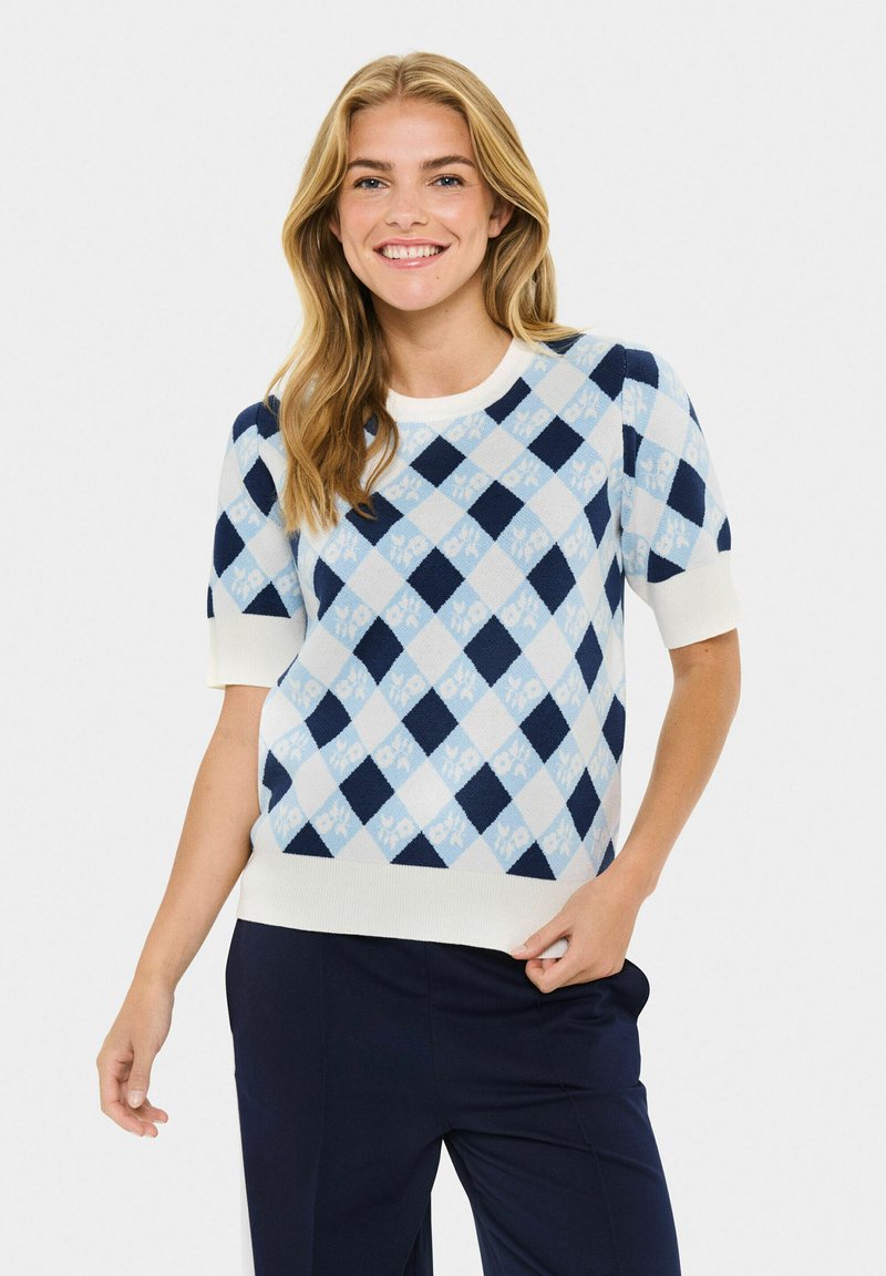 Mujer sonriente con cabello largo y rubio, vestida con un suéter de manga corta de patrón argyle en azul y blanco y pantalones oscuros.