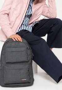Gray Eastpak backpack with textured fabric, two front pockets, and logo patch. Features drawstring details and a rounded shape.