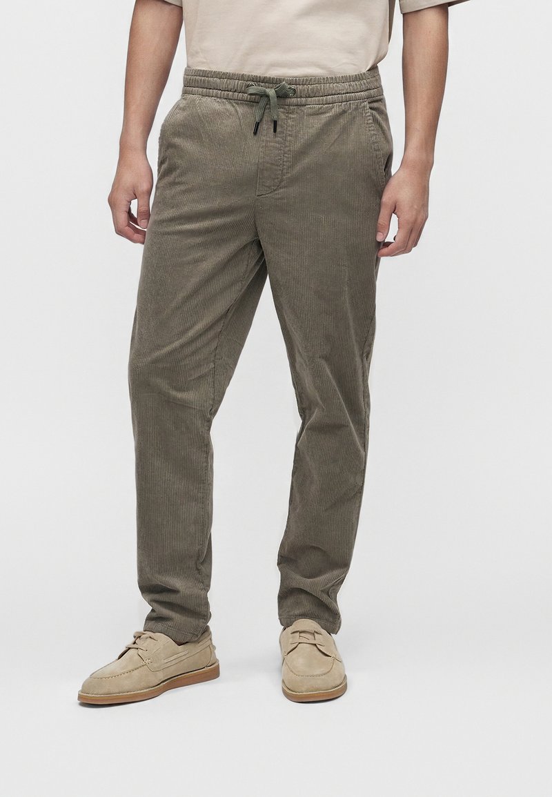 Uomo che indossa pantaloni di velluto a coste verde oliva con vita regolabile a coulisse e mocassini beige in suede, in piedi davanti a uno sfondo semplice.
