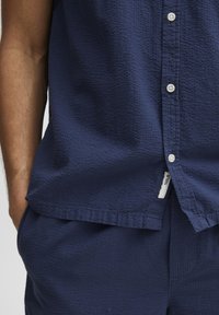 Camisa de manga corta azul marino hecha de un material texturizado, con un cierre frontal de botones y una etiqueta de marca visible en el dobladillo.