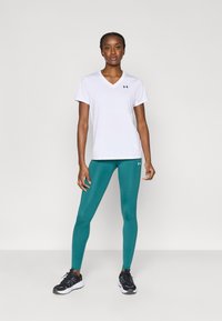 Baltas sportinis v formos marškinėliai trumpomis rankovėmis, derinami su žalsvais tamprėmis. Juodi sportbačiai su akcentais, šviesiai pilkas fonas.