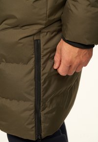 Olivgrüne Steppjacke mit wattierter Textur, ausgestattet mit einer seitlichen Reißverschlusstasche und schwarzem Reißverschluss. Die Ärmelbündchen sind mit schwarzem Besatz versehen.