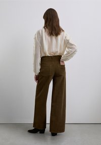 Pantalon large en velours côtelé marron à taille haute avec poches arrière, associé à un chemisier léger et transparent à manches longues, vu de dos.