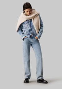 Chaqueta de mezclilla azul claro sobre una suave bufanda de lana beige, combinada con jeans rectos azul claro y zapatos negros. Textura suave y diseño casual.